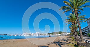 Abandoned sailboat on the beach. Rows of palm trees line water`s edge in Ibiza, St Antoni de Portmany Balearic Islands, Spain.