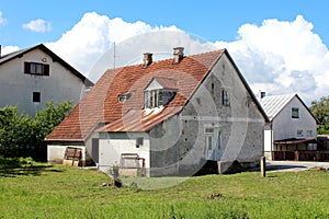 Abandoned small old suburban family house with dilapidated damaged white facade and red roof tiles mixed with rusted metal tiles