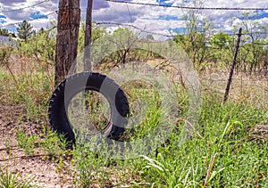 An abandoned truck tire leans against a wooden fence post in a scrubby landscape.