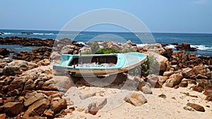 Abandoned turquoise boat filled with plastic bottles and rubbish on sandy shore. Ocean waves crash on rocky beach with