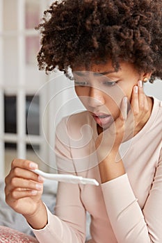 Abortion problem concept. Puzzled black woman holds pregnancy test, feels depressed and sad, poses at home, keeps hand