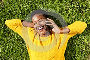 Above of happy young black woman lying on green grass and talking with mobile phone