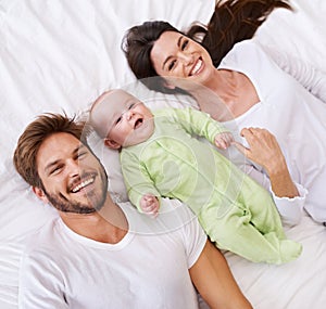 Above, portrait and happy parents with baby on bed for love, care and quality time together at home. Smile of mother