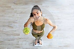 Above view of happy Indian lady holding citrus fruit and cabbage, standing on scales, choosing healthy diet, full length