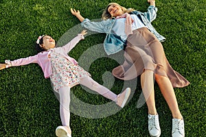 Above view image of happy mom lying on the green grass with her cute little girl in the park. Cheerful young woman relaxing with