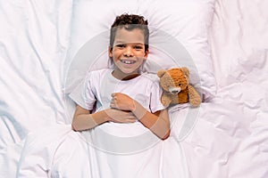 Above view portrait of positive adorable funky schoolkid lay comfy bed pillow blanket favorite soft teddy bear toy