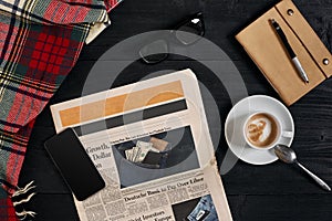 Above view of Smart phone, newspaper, scarf in a cage, glasses with notebook and cup of latte coffee on black wooden