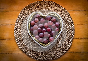 Above view of the white heart-shaped plate full of red grapes, resting on a straw mat. Wooden background