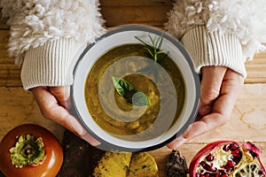 Above view of woman hands holding cream vegetable soup on a wooden table with autumn decorations leaves and fruits. Healthy winter
