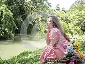Absent minded asian woman in pink dress with a bouquet of flowers sitting in the garden near canal.