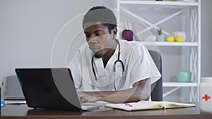 Absorbed African American man typing on laptop keyboard sitting in hospital office indoors. Concentrated serious doctor