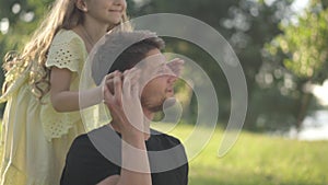 Absorbed Caucasian man sitting on park lawn surfing Internet as cheerful girl covering eyes with hands laughing