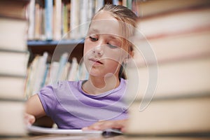 Absorbed by a good story. A cute young girl doing schoolwork while surrounded by books at the library.