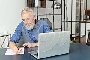 Absorbed pensive senior businessman in smart casual shirt sits at the desk