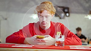 Absorbed redhead young man using smartphone sitting at bar counter with cocktail glass as man passing at background