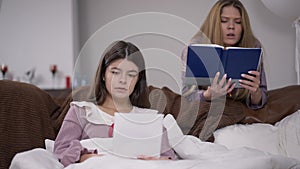 Absorbed young woman sitting on couch in dorm room as blurred friend reading at background. Two concentrated Caucasian