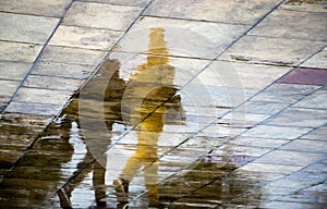 Abstract blurry silhouette reflections of unrecognizable people walking under umbrella on wet city street pavement on a rainy day