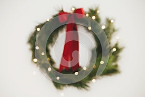 Abstract Christmas background. Blurred stylish christmas wreath with red bow and lights on white wall. Merry Christmas and Happy