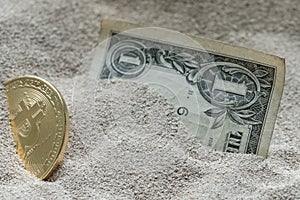 Bitcoin currency seen partially buried in Silicon Sand together with a One Dollar Banknote.