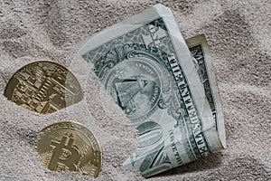 Bitcoin currency seen partially buried in Silicon Sand together with a One Dollar Banknote.