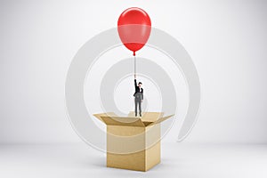 Abstract image of young european businessman flying on red balloon out of paper box on light background. Success, business and