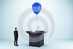 Abstract image of young european businessman looking at flying blue balloon out of black paper box on light background. Success,