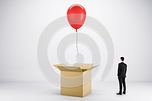 Abstract image of young european businessman looking at flying red balloon out of paper box on light background. Success, business