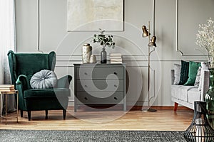 Abstract painting on grey wall o contemporary living room interior with emerald green armchair with round pillow, commode and