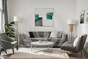 Abstract painting on grey wall o contemporary living room interior with emerald green armchair with round pillow, commode and couc