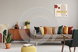 Abstract painting on grey wall of retro living room interior with beige sofa with pillows, vintage dark green armchair and yellow