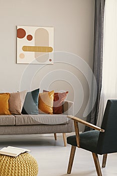 Abstract painting on grey wall of retro living room interior with beige sofa with pillows, vintage dark green armchair and yellow