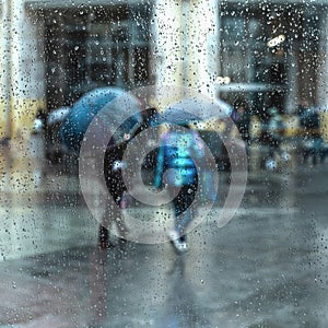 Abstract people under the umbrella, rainy weather, blurred motion. View through glass window with rain drops on glass