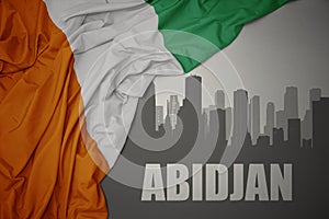 Abstract silhouette of the city with text Abidjan near waving colorful national flag of cote divoire on a gray background