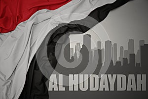 Abstract silhouette of the city with text Al Hudaydah near waving national flag of yemen on a gray background