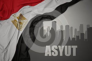 Abstract silhouette of the city with text Asyut near waving colorful national flag of egypt on a gray background