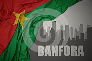 Abstract silhouette of the city with text banfora near waving colorful national flag of burkina faso on a gray background