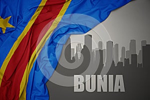 Abstract silhouette of the city with text Bunia near waving colorful national flag of democratic republic of the congo on a gray