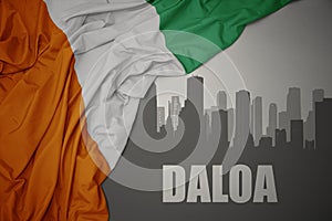Abstract silhouette of the city with text Daloa near waving colorful national flag of cote divoire on a gray background