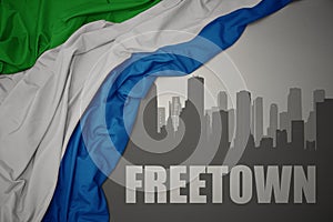 Abstract silhouette of the city with text Freetown near waving colorful national flag of sierra leone on a gray background