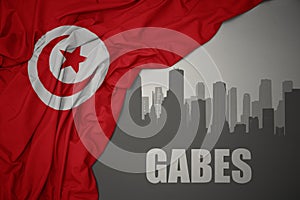 Abstract silhouette of the city with text Gabes near waving colorful national flag of tunisia on a gray background