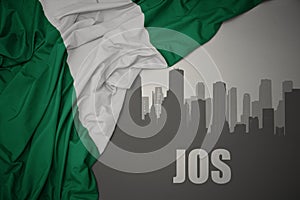 Abstract silhouette of the city with text Jos near waving colorful national flag of nigeria on a gray background