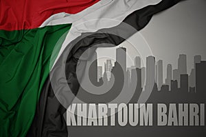 Abstract silhouette of the city with text Khartoum Bahri near waving colorful national flag of sudan on a gray background