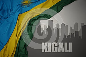 Abstract silhouette of the city with text Kigali near waving colorful national flag of rwanda on a gray background