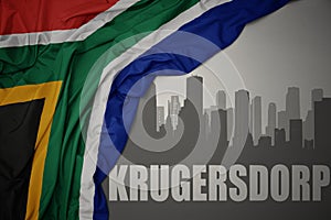 Abstract silhouette of the city with text Krugersdorp near waving colorful national flag of south africa on a gray background