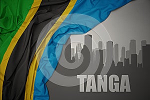 Abstract silhouette of the city with text Tanga near waving colorful national flag of tanzania on a gray background