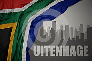 Abstract silhouette of the city with text Uitenhage near waving colorful national flag of south africa on a gray background