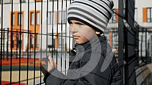 Abused and stressed little boy holding metal fence and looking on children palyground