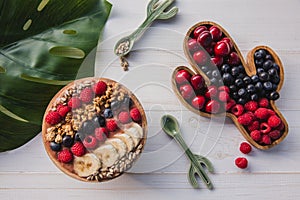 Acai smoothie, granola, seeds, fresh fruits in a wooden bowl with cactus spoon. Plate filled with berries on white