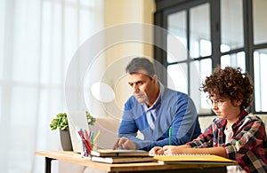 Achieving knowledge together. Concentrated teenaged hispanic boy in glasses making notes while sitting at the desk