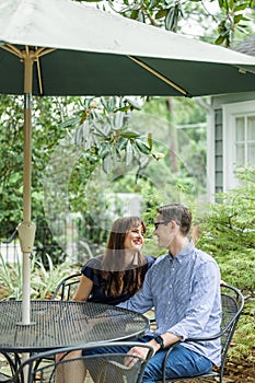 Acouple of a husband and wife sitting under a shaded umbrella patio table outside in the backyard